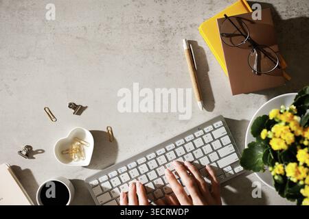 Ein femininer Arbeitsbereich mit Händen auf der Tastatur, einer Tasse Kaffee, gelben Blumen und Schreibwaren. Helles Morgenlicht leuchtet das saubere A auf Stockfoto