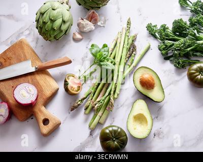 Verschiedene Gemüsesorten werden auf einer Marmorarbeitsfläche in einer Küche platziert. Ein Messer und ein Schneidebrett sind in der Nähe. Frische Früchte und Kräuter tragen zum gesunden i bei Stockfoto