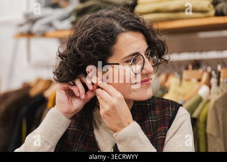 Eine reife Frau mit lockigem Haar und Brille lächelt, während sie ihre Ohrringe in einer stilvollen, urbanen Boutique anpasst. Sie verkörpert den modernen Stadtstil und cas Stockfoto
