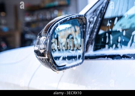 Nahaufnahme eines modernen Autospiegels, der während einer professionellen Autowäsche mit Seifenschaum und Wassertropfen bedeckt ist, mit verschwommenem Garagenhintergrund und Clea Stockfoto