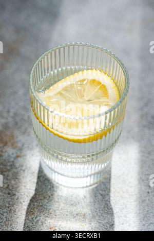 Erfrischendes Glas Zitronenwasser mit Zitrusscheibe in einem gerippten Becher, auf einer grauen Steinplatte mit weichem natürlichem Sonnenlicht und Schatten, minimal Stockfoto