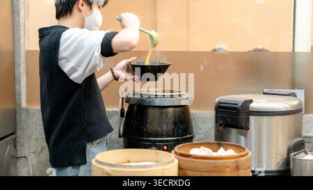 Ein Koch gießt heiße Suppe in eine Schüssel in einer taiwanesischen Restaurantküche, umgeben von Dampfern, die mit frischen Knödeln gefüllt sind. Die Umgebung strahlt ein warmes, au aus Stockfoto