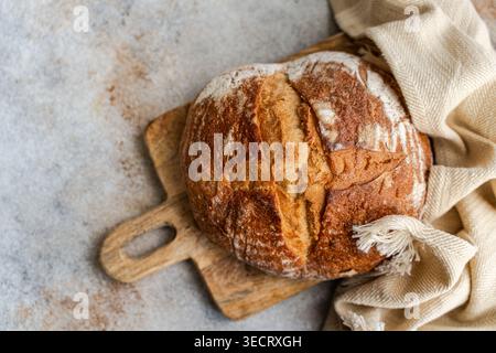 Frisch gebackenes Sauerteigbrotbrot mit goldener Kruste und mehlbestäubter Ritzel, auf einem hölzernen Schneidebrett neben einem Leinentuch auf einer steinplatte Stockfoto