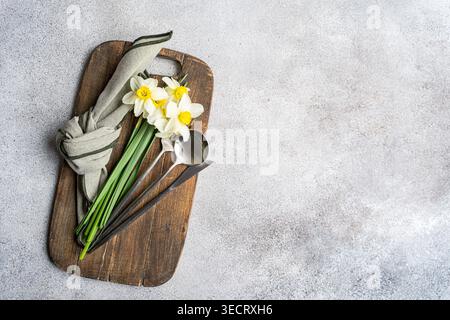 Frischer Frühlings-Narzissen-Bouquet, in Leinen auf einem rustikalen Holzbrett mit Metalllöffel gewickelt, von oben auf einem hellen strukturierten Hintergrund geschossen. Stockfoto