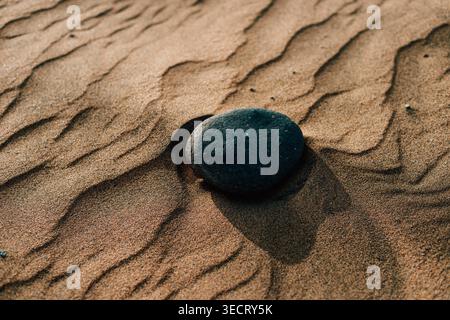 Nahaufnahme eines glatten vulkanischen Steins, der bei Sonnenuntergang in den Dünen von Maspalomas auf Gran Canaria auf windgeformtem, gewelltem Sand ruht und die ruhige, minimale Wüste einfängt Stockfoto