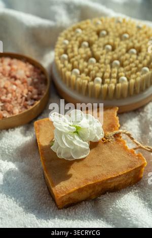 Warmes Wellness-Stillleben auf einem weichen Handtuch mit einer hölzernen Spa-Bürste, einer Schüssel mit rosa Spa-Salz und handgemachtem Seifenbügel mit weißer Blume, ruhiger Wellness-Stimmung Stockfoto