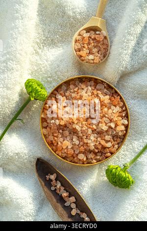 Nahaufnahme rosafarbener Spa-Salzkristalle in einer Schüssel auf einem weichen weißen Handtuch, dekoriert mit einer hölzernen Schaufel und frischen grünen Blumen für eine ruhige Wellness-Spa-Umgebung Stockfoto