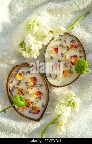 Zwei Spa-Kokoskerzen in Halbschalen, verziert mit getrockneten Rosenblättern und Lavendel, angeordnet auf einem weichen Handtuch mit frischen weißen Blumen für eine ruhige Atmosphäre Stockfoto