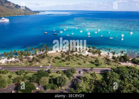 Blick aus der Vogelperspektive auf einen von Palmen gesäumten Strand und eine türkisfarbene Lagune mit Segelbooten vor Anker, Korallenriffmuster und Küstenstraße auf einer tropischen Insel auf Französisch Stockfoto