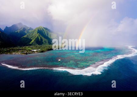 Aus der Vogelperspektive auf eine tropische Insellagune mit türkisfarbenem Riffwasser, brechenden Wellen und einem lebhaften Regenbogen über nebeligen grünen Bergen in Französisch-Polynesien Stockfoto