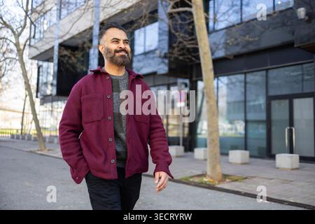 Arabischer Mann mit Bart, der selbstbewusst auf einer Stadtstraße neben einem modernen Bürogebäude läuft, an einem kühlen Tag eine burgunderrote Jacke trägt, urbanen Lebensstil und Stockfoto