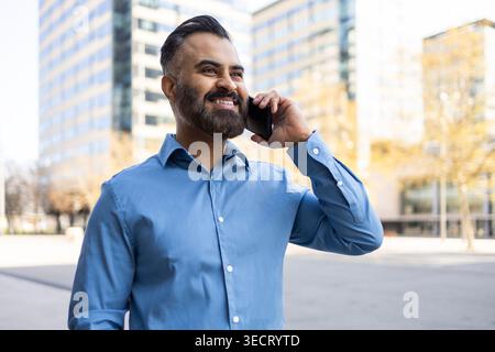 Ein lächelnder arabischer Geschäftsmann in blauem Hemd spricht draußen auf einem Smartphone auf einer modernen Stadtstraße mit Bürogebäuden und fängt selbstbewusste professionelle Mitarbeiter ein Stockfoto