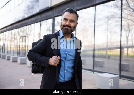 Ein selbstbewusster arabischer Geschäftsmann im Anzug geht vor ein modernes Bürogebäude aus Glas, trägt einen Rucksack und blickt zur Seite und vermittelt Führung A Stockfoto