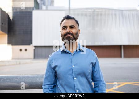 Selbstbewusster arabischer Geschäftsmann in blauem Hemd, der draußen an einem modernen Bürogebäude steht und mit einem ruhigen Lächeln nach vorne blickt; professionelles Porträt mit urba Stockfoto