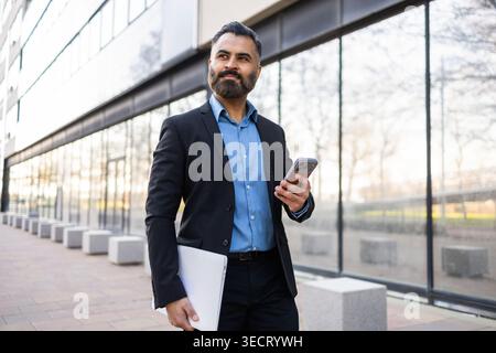 Ein selbstbewusster arabischer Geschäftsmann im Anzug geht vor einem modernen Bürogebäude, hält ein Smartphone und Laptop, konzentriert und bereit für Arbeit, Führung, Stockfoto