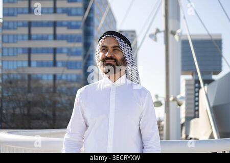Ein lächelnder arabischer Geschäftsmann in traditioneller sheikh-Kleidung steht draußen in einem modernen Stadtviertel und vermittelt Vertrauen, Führungsstärke und Erfolg in Stockfoto