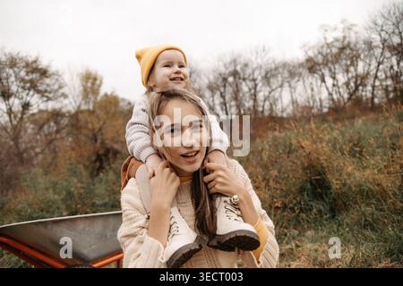 Fröhliche Schwestern genießen einen Huckepack-Ritt in einem Herbstpark mit kuscheligen Strickpullis und warmer Mütze. Natürliches Außenporträt mit Bäumen, Laub und Familie Stockfoto