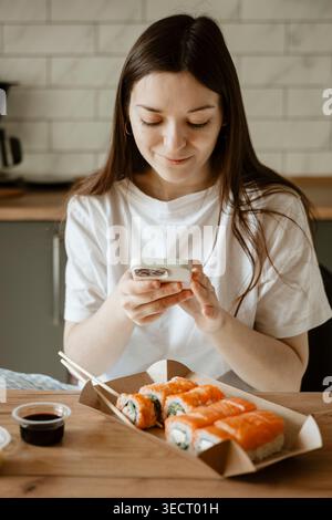 Eine Frau genießt Sushi zu Hause an ihrem freien Tag und hält den Moment mit ihrem Smartphone fest. Die Szene vermittelt Entspannung und kulinarische Genüsse in einem gemütlichen Set Stockfoto