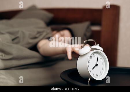 Eine Person genießt einen faulen Tag, indem sie den Wecker ausschaltet, während sie bequem im Bett liegt, und veranschaulicht das Konzept der Bettenverrottung. Stockfoto
