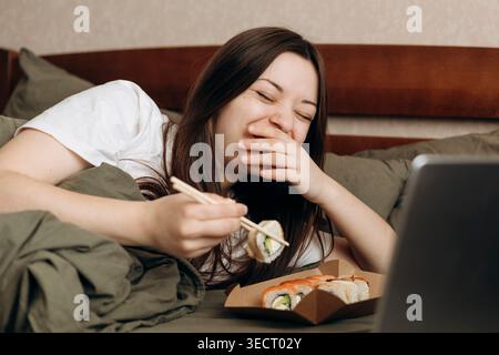 Eine Frau genießt einen entspannten Tag zu Hause, lacht beim Sushi-Essen im Bett mit Essstäbchen. Die gemütliche Umgebung und der freudige Moment betonen eine Perfec Stockfoto