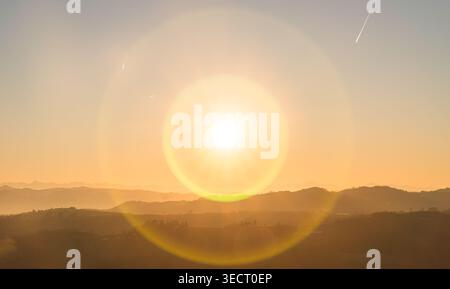 Warmer Sonnenuntergang über den sanften Hügeln des Piemonte, Norditalien, mit trüben Bergsilhouetten und goldenem Himmel, die eine ruhige Landschaft in der Abenddämmerung einfangen. Stockfoto