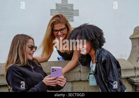 Drei junge Freundinnen treffen sich im Freien, lächeln und lachen, während sie sich einen Smartphone-Bildschirm teilen, die zwanglose soziale Zeit und moderne Kommunikation genießen Stockfoto