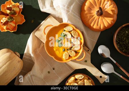 Eine lebhafte Kürbissuppe im Herbst, garniert mit Croutons, frischen Kräutern und Samen, serviert in einer kürbisförmigen Schüssel auf dunkelgrünem Hintergrund. Perfekt für Stockfoto