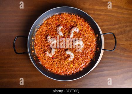 Blick von oben auf traditionellen spanischen Reis mit Meeresfrüchten, gekocht in einer Paella-Pfanne, mit Garnelen auf reichhaltigem Tomaten-Safranreis, serviert auf einem rustikalen Holztisch, Hausstraße Stockfoto