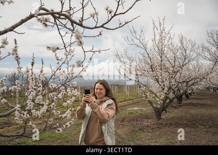 Eine Frau macht mit ihrem Smartphone ein Foto zwischen blühenden Aprikosenbäumen, mit dem schneebedeckten Berg Ararat in der Ferne im ländlichen Armenien duri Stockfoto