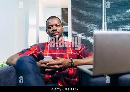 Selbstbewusster junger Schwarzer Mann, der sich auf einem Sofa in einem modernen Büro entspannt und ein Smartphone neben einem offenen Laptop hält. Lässiges Business-Porträt mit natürlichem Long Stockfoto