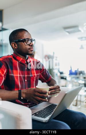 Ein durchdachter, professioneller Mann in Brille sitzt auf einem Sofa in einem modernen Büro, hält eine Kaffeetasse, während er an einem Laptop arbeitet und reflektiert über Strategie und p Stockfoto