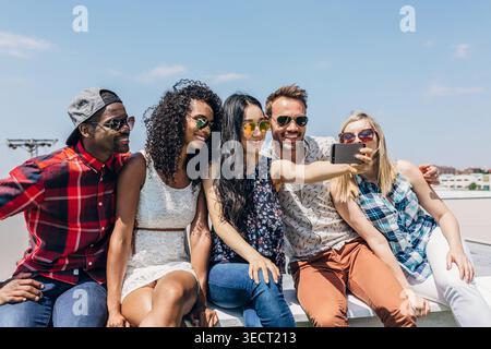 Verschiedene Gruppen junger Freunde sitzen zusammen auf einem Dach, lächeln und machen an einem hellen Sommertag ein Selfie mit einem Smartphone, und tragen sich lässig aus Stockfoto