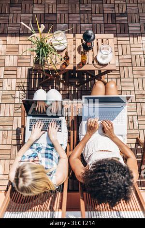 Draufsicht auf zwei Frauen, die draußen auf einer sonnigen Terrasse an Laptops arbeiten, mit Kaffee, Getränken und Pflanzen auf einem kleinen Tisch, Aufnahmen von Fernarbeit, Teamarbeit A Stockfoto