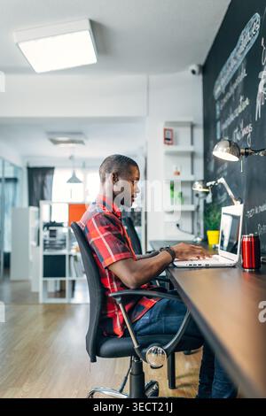 Seitenansicht eines fokussierten jungen professionellen Schreibtischers auf einem Laptop an einem Schreibtisch in einem modernen Büro, mit Wandnotizen an der Tafel, Lampenbeleuchtung und kreativer Arbeit Stockfoto