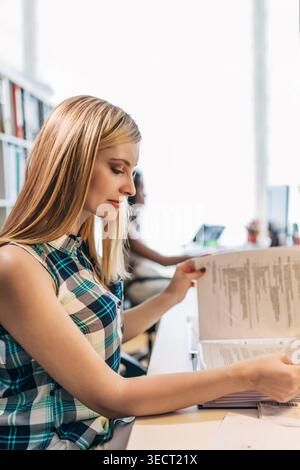 Fokussierte junge professionelle Frau, die gedruckte Dokumente an einem Schreibtisch in einem hellen, modernen Büro mit Akten und Laptop in der Nähe prüft, was Produktivität fördert Stockfoto