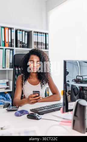 Junge professionelle Frau in einem hellen, modernen Büro, am Schreibtisch sitzt und Smartphone neben Computermonitor, Tastatur und Dateien überprüft, konzentriert sich auf W Stockfoto