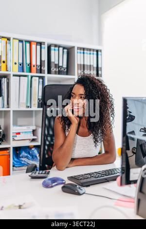 Lächelnde junge Geschäftsfrau, die an einem modernen Schreibtisch sitzt und nachdenklich beiseite blickt. Computermonitor, Tastatur, Taschenrechner und Regale mit Ordnern Stockfoto