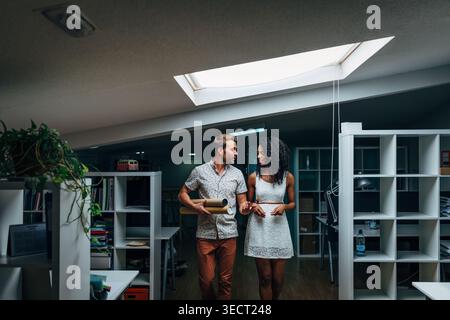 Ein junges Paar spaziert durch ein modernes Loft-Apartment unter einem Oberlicht, diskutiert Renovierungspläne und hält einen gerollten Plan, umgeben von Regalen Stockfoto