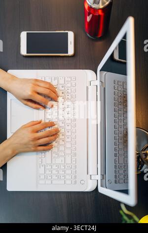 Draufsicht der Hände, die auf einem weißen Laptop an einem dunklen Holzschreibtisch tippen, mit Smartphone und einer roten Getränkedose in der Nähe, die einen produktiven Arbeitsplatz für die Wiederaufnahme zeigt Stockfoto