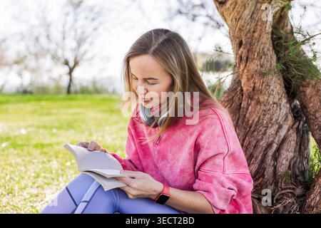 Eine Frau sitzt unter einem Baum in einem ruhigen Park, tief in ein Buch vertieft. Sie trägt ein rosafarbenes Sweatshirt und Kopfhörer um den Hals und genießt einen Quie Stockfoto