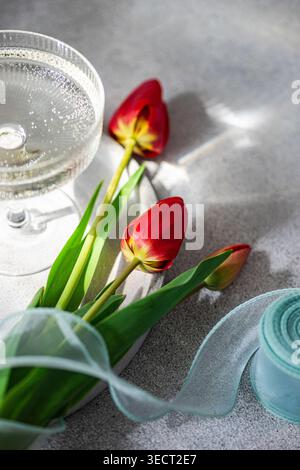 Elegantes Stilleben aus roten Tulpen auf einem strukturierten grauen Tisch mit transparentem Band und funkelndem Coupé-Glas bei weichem Tageslicht mit sanftem Schatten Stockfoto