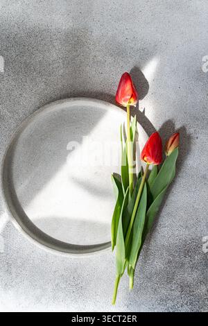 Drei rote Tulpen mit grünen Blättern ruhen auf einer weißen Keramikplatte auf einem grauen Betontisch, beleuchtet von natürlichem Sonnenlicht und weichem Schatten; minimale Stilllebensdauer Stockfoto