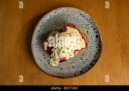 Blick von oben auf ein überdachtes Frühstück mit Eiern Benedict mit Hollandaise-Sauce und Schnittlauch auf einem rustikalen Keramikteller, serviert auf einem Holztisch in warmen More Stockfoto