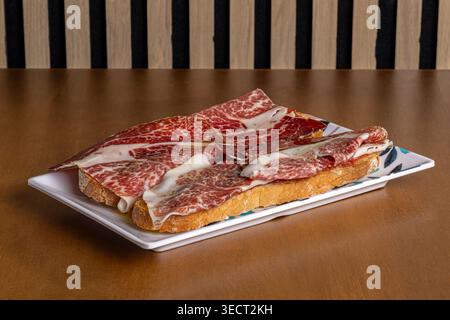 Morgens Frühstück auf einem rechteckigen Teller mit geröstetem Brot, garniert mit dünnen Scheiben Räucherschinken, serviert auf einem Tisch mit einem einfachen gestreiften Backgro Stockfoto