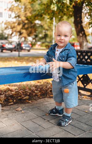 Niedliches Kleinkind in Jeansjacke und Shorts steht neben einer blauen Bank in einem sonnigen Herbstpark und lächelt mit verschwommenen Bäumen und einer Straße in der Badewanne in die Kamera Stockfoto