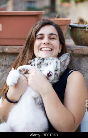 Eine glückliche junge Frau umarmt einen flauschigen Collie-Welpen draußen und lächelt in die Kamera. Warmes Lifestyle-Porträt, das Zuneigung, Gesellschaft und zeigt Stockfoto