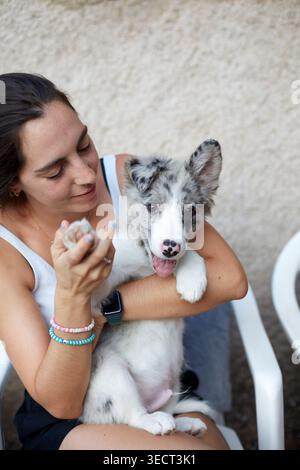 Junge Frau hält und kuschelt einen blauen merle Border Collie Welpen, lächelt beim Training oder spielt mit ihrem Hund auf einer sonnigen Terrasse im Freien. Stockfoto