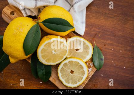 Frische Zitronen und in Scheiben geschnittene Zitronenrunden mit grünen Blättern auf einem rustikalen Holzschneidebrett neben einem Küchentuch, das natürliche Zitrus-fru zeigt Stockfoto