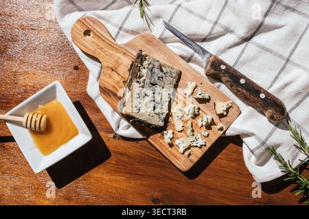 Rustikale Essensszene mit Blauschimmelkäse auf einem hölzernen Schneidebrett, zerbröckelten Stücken, einem Vintage-Messer, Rosmarinzweigen und einer Schüssel Honig auf einem Tex Stockfoto