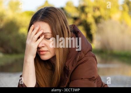 Eine besinnliche Frau mit langen braunen Haaren sitzt draußen, ihre Hand liegt sanft auf ihrem Gesicht. Sie scheint tief in Gedanken zu sein, umgeben von friedlichen Stockfoto